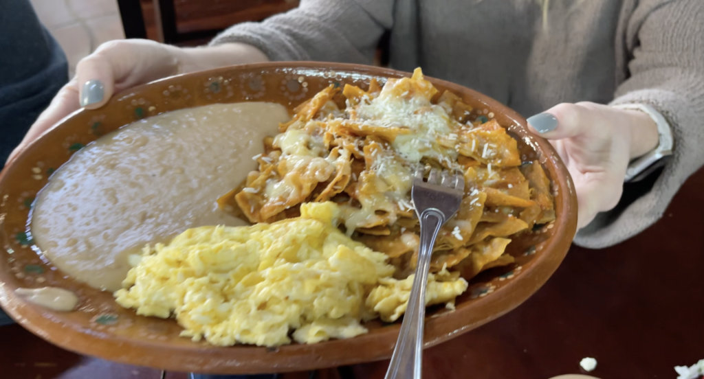 A plate of chilaquiles, a traditional Mexican dish made with fried tortilla chips simmered in salsa and topped with cheese, onions, and crema. It is served alongside refried beans and scrambled eggs. This dish is from La Cocina de Dona Esthela, a popular restaurant in the Valle de Guadalupe. Valle de Guadalupe Mexico Tour.