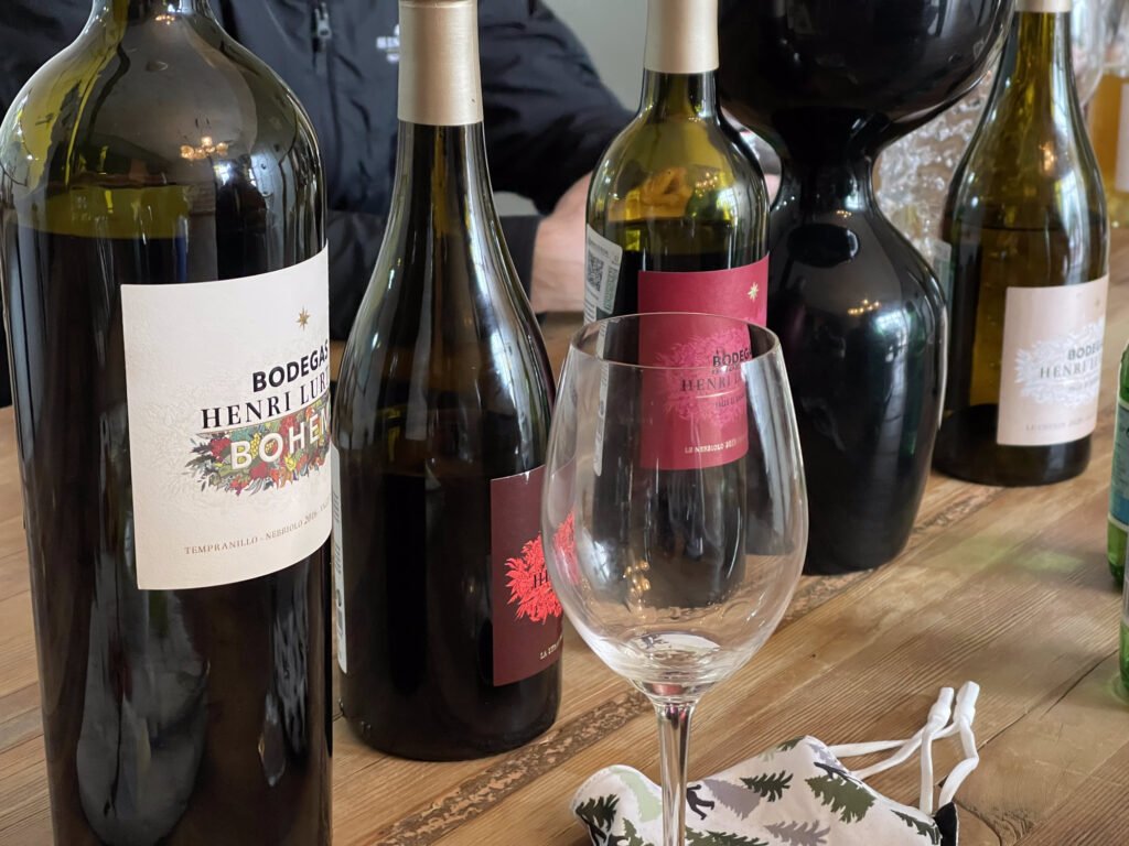 A variety of wine bottles from Bodega Henri Lurton lined up on a wooden table. A wine glass sits in front of the bottles, and a face mask with a tree pattern rests on the table. Wine tasting at Henri Lurton, Valle de Guadalupe Mexico Tour.