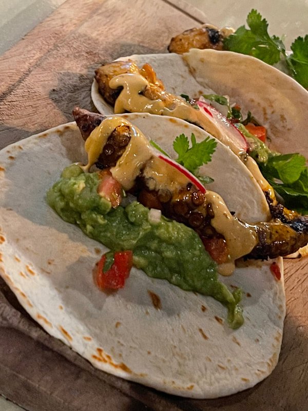 Two tacos filled with grilled octopus tentacles, topped with creamy sauce, cilantro, and radishes on a wooden board. A portion of guacamole with diced tomatoes sits next to one taco.