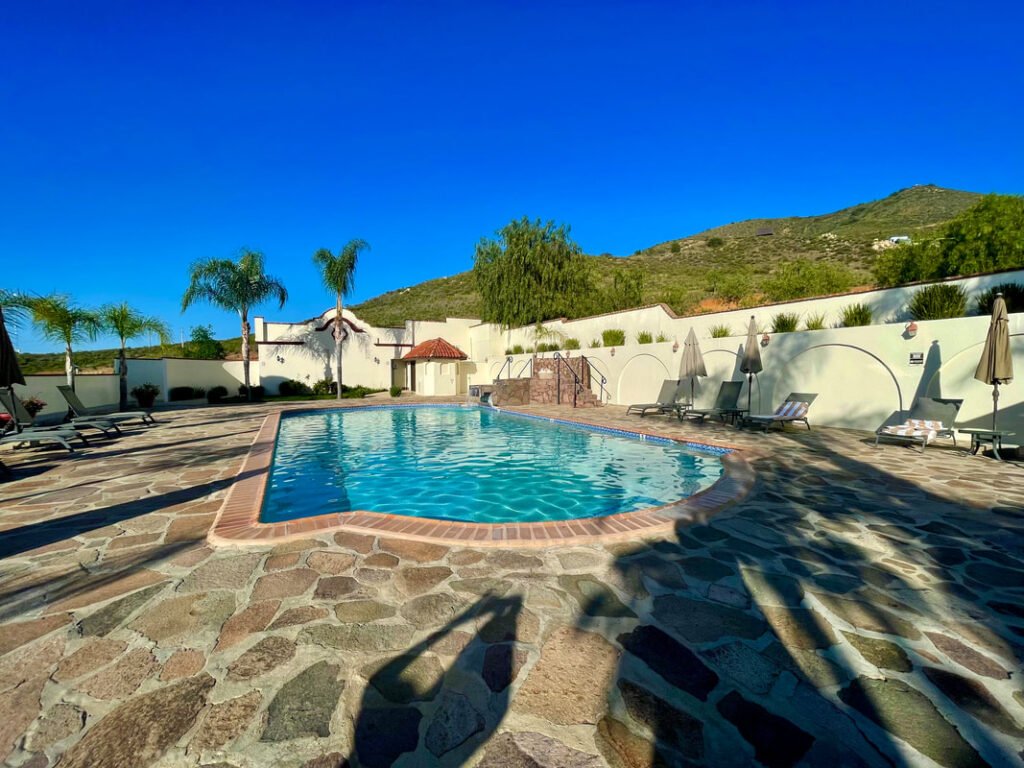 Outdoor swimming pool area with clear blue water, stone-tiled deck, lounge chairs, umbrellas, palm trees, and hills in the background. Mexico wine tours.