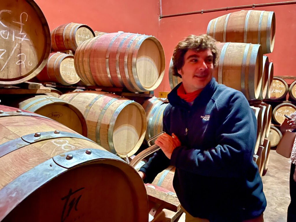 Person standing in a room filled with wooden barrels, likely used for aging wine. The person is interacting with one of the barrels. Mexico wine tours.