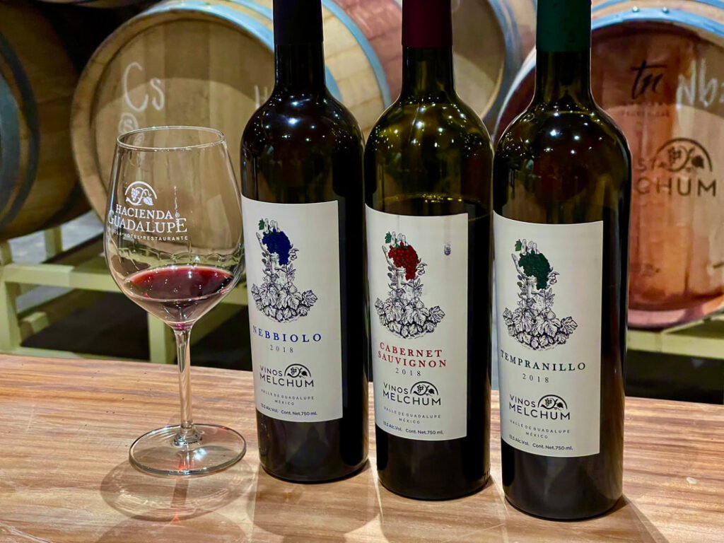 "Three bottles of wine labeled 'Nebbiolo 2018,' 'Cabernet Sauvignon 2018,' and 'Tempranillo 2018' from 'Vinos Melchum' in Valle de Guadalupe, alongside a wine glass with 'Hacienda Guadalupe Restaurante' text, capturing the diverse offerings of Mexico Wine Tours and VIP Winery Vacations.