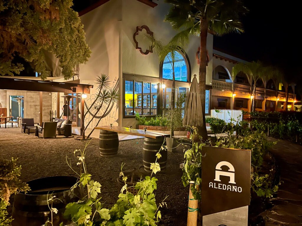 Outdoor seating area of a restaurant named "Aledaño," with wooden barrels as tables, warm lighting, palm trees, and decorative elements, showcasing the inviting atmosphere of Mexico Wine Tours and VIP Winery Vacations.