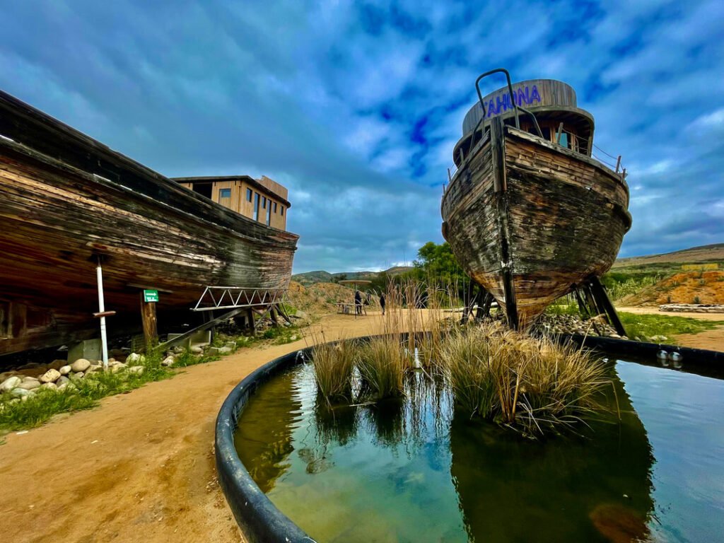 Old, weathered wooden boats on land, with one named "AHANA" and surrounded by a dry, rocky landscape and a small pond, are part of an outdoor display showcasing the unique experiences offered by Mexico Wine Tours and VIP Winery Vacations.