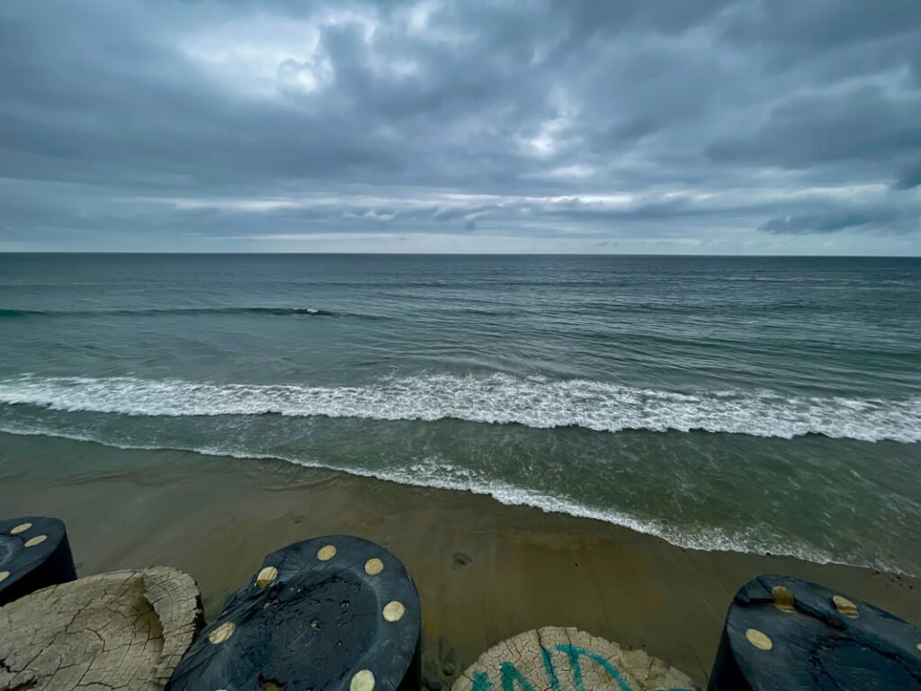 "View of the ocean under a cloudy sky, with gentle waves rolling onto a sandy beach and circular concrete structures with black surfaces and yellow circular markings in the foreground.