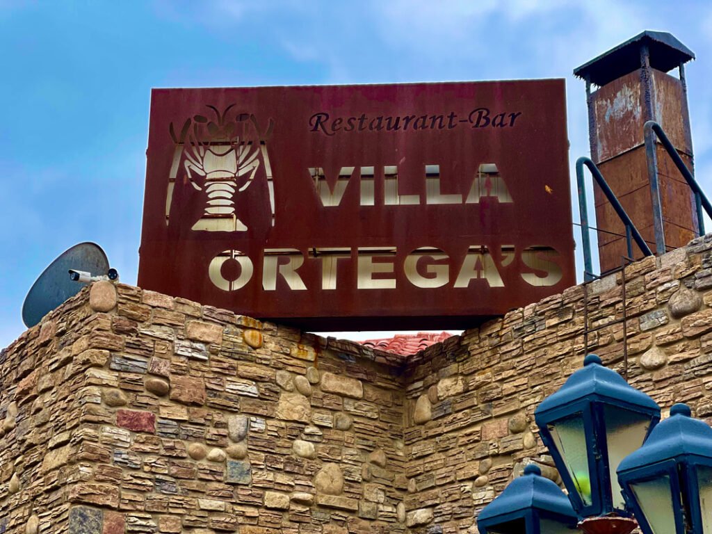 The sign for "Restaurant-Bar Villa Ortega's" stands prominently on a stone wall, with a lobster graphic and cut-out letters illuminated by the sunlight, representing the inviting atmosphere of Mexico wine tours.