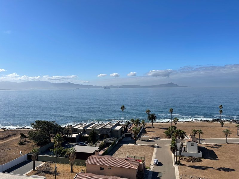 A coastal scene with clear blue sky, calm ocean waters, buildings and palm trees in the foreground, a rocky shoreline, and distant mountains and islands.