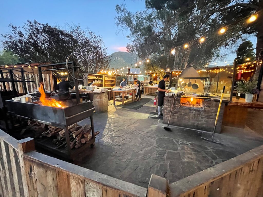 An outdoor cooking area features multiple grills with open flames, various cooking utensils, and string lights overhead, capturing the rustic ambiance of Mexico wine tours.