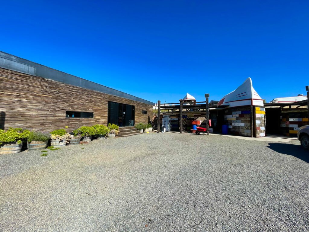 A modern building with wooden exterior, surrounded by potted plants and gravel, stands next to a vibrant, pointed-roof structure showcasing the unique charm of Mexico wine tours.