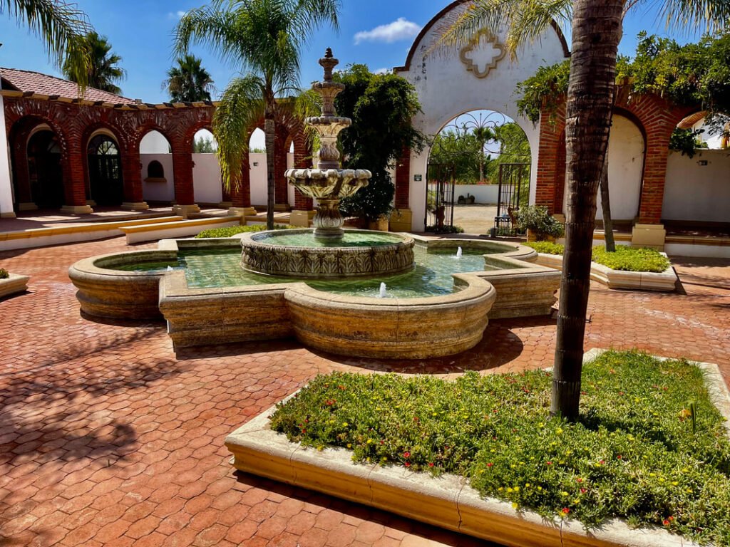 Courtyard with a central multi-tiered fountain, surrounded by a shallow pool with small water jets. The courtyard is paved with red bricks, with several palm trees and green plants in rectangular planters. In the background, arched structures made of red bricks with white walls. Clear blue sky. Mexico wine tours.