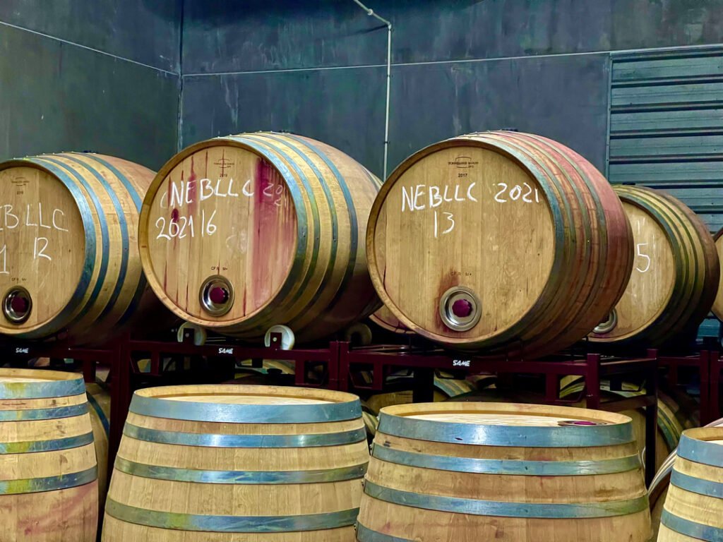 A collection of wooden barrels, likely used for aging wine, arranged in a storage area. Each barrel has text indicating contents and dates, like "NEBLLC 2021." The setting appears to be a winery storage room. Mexico wine tours.