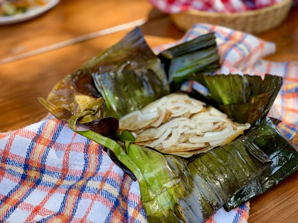 Dish wrapped in banana leaves on a checkered cloth, containing sliced onions.