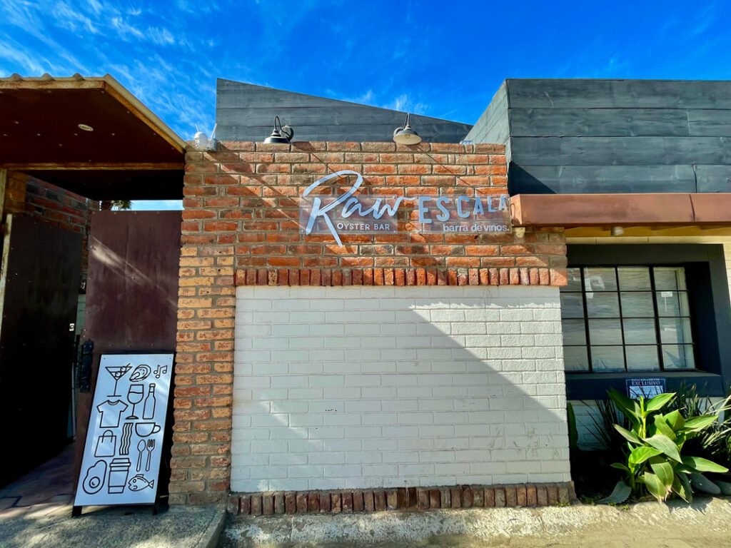 Exterior of "Raw ESCALA Oyster Bar barra de vinos" building with a brick and white-painted wall, clear blue sky, and some plants visible.