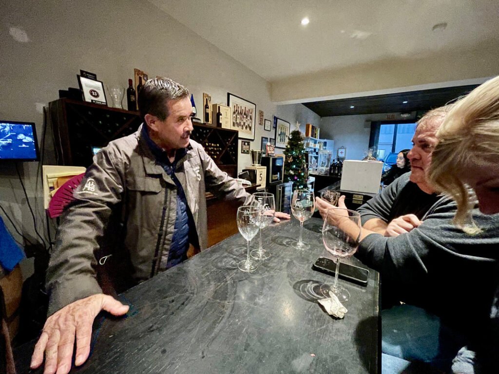 Three people standing around a bar counter with wine glasses, a smartphone, and a decorated Christmas tree in the background.