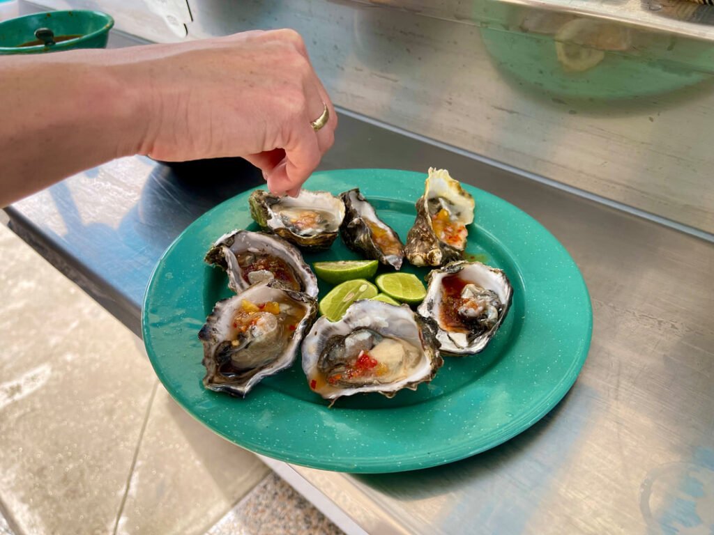 A hand reaching for a plate of six opened oysters with sauce or seasoning, accompanied by three lime wedges on a green plate, set on a metallic surface