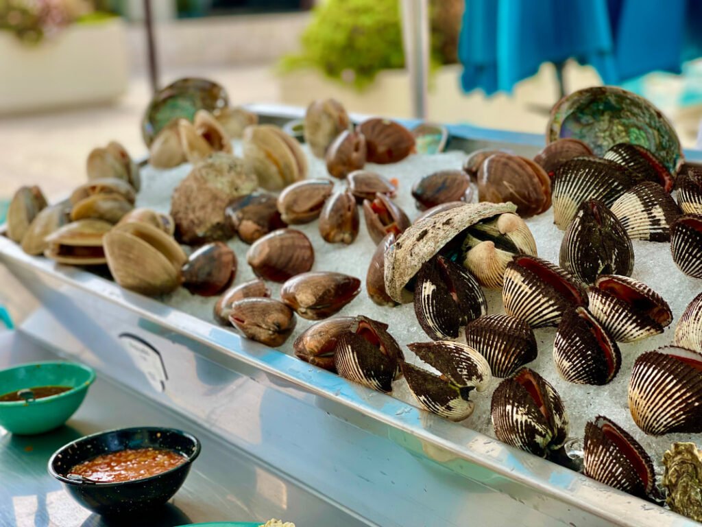 A display of various types of shellfish on a bed of ice, including clams and oysters. Two bowls of sauce or condiments are placed in front of the shellfish display, arranged to showcase their variety and freshness.