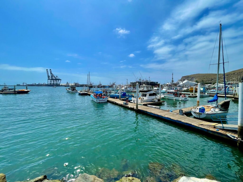 A bustling marina with various boats docked at wooden piers, clear blue-green water, and a few scattered clouds in the sky. Large industrial cranes are visible in the background, suggesting a nearby port.