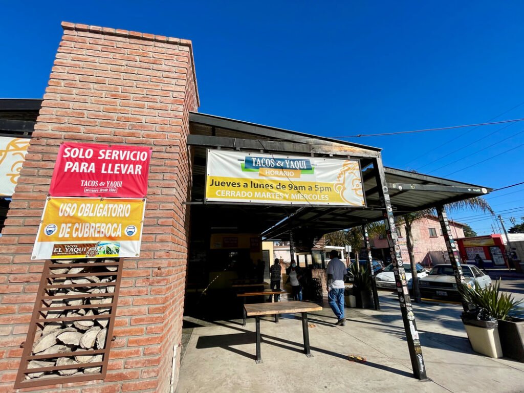 The exterior of the taco restaurant "Tacos & Yaqui" with a brick facade and signs indicating "TAKEOUT SERVICE ONLY" and "MANDATORY USE OF FACE MASK." The restaurant's hours are also displayed as "Thursday to Monday from 9am to 5pm, CLOSED TUESDAY AND WEDNESDAY."