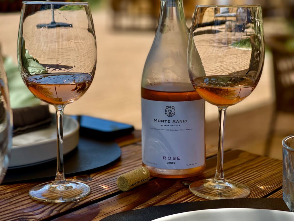 A bottle of Monte Xanic Rosé wine from 2020, alongside two glasses of the vibrant rosé, set against the serene backdrop of an outdoor Mexico wine tour.